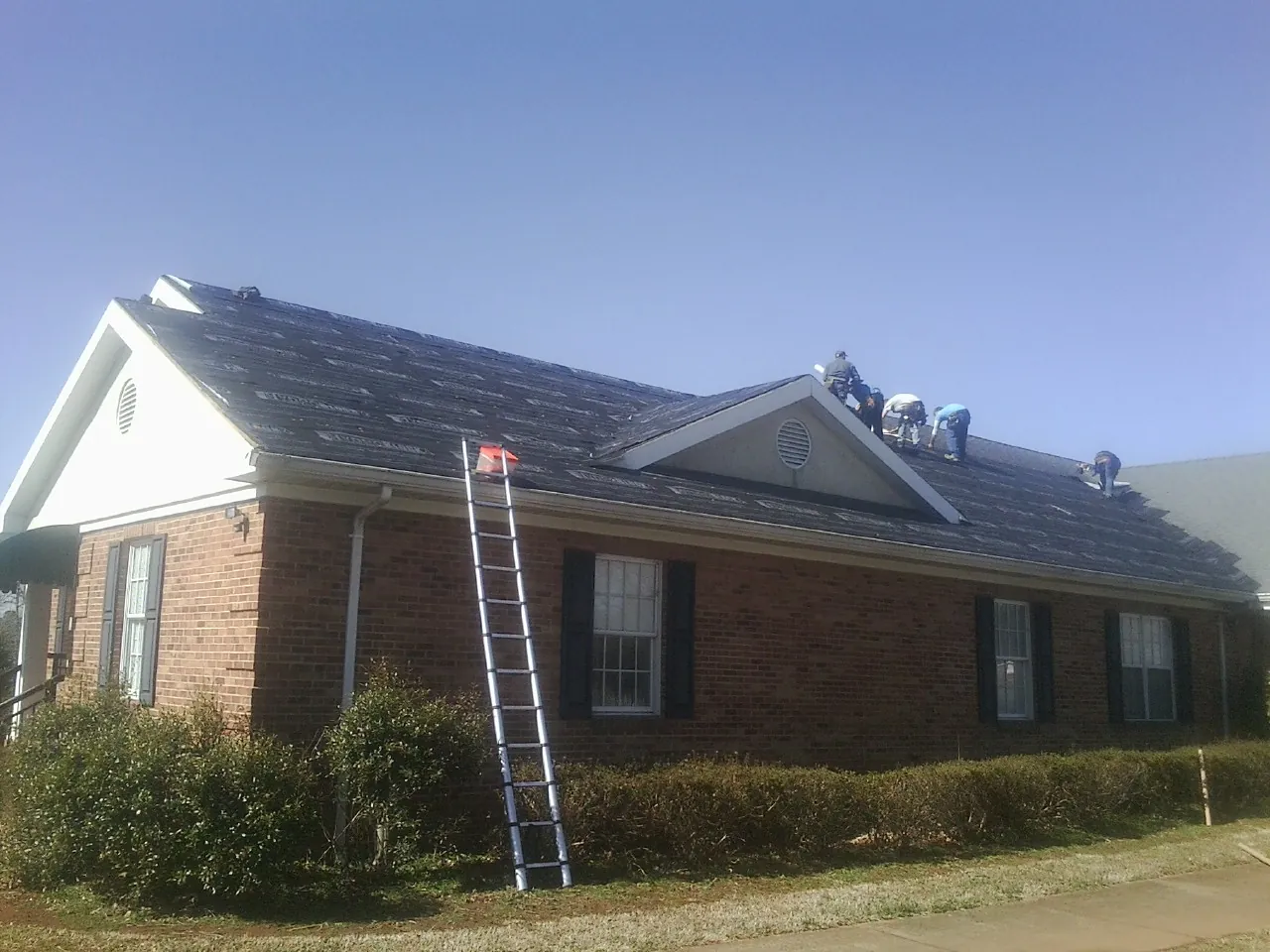 Crew tearing shingles off a brick ranch roof with a ladder set — Emergency Roof Repair work by our crew in Center Ossipee