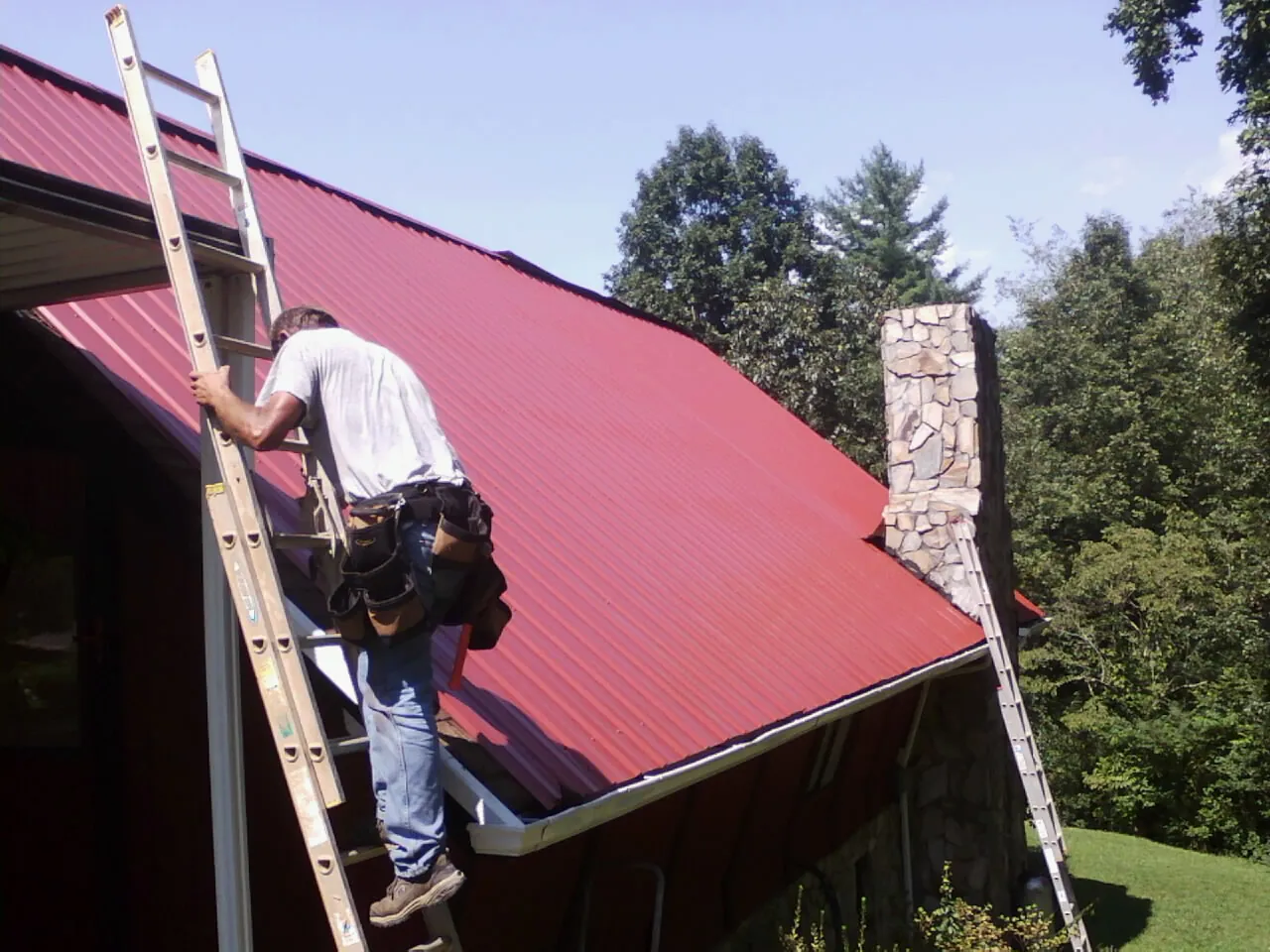 Roofer on a ladder beside a red metal roof and stone chimney — Soffit & Fascia Repair by Roofing Brothers in Center Ossipee