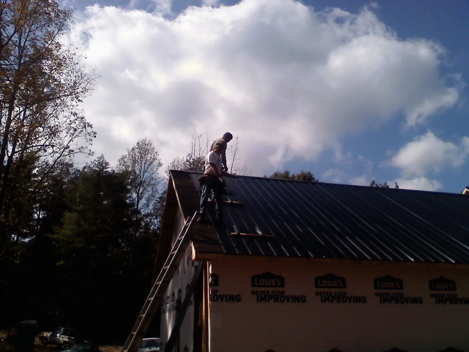Roofer fastening dark metal panels high against a cloudy sky, from a Roof Inspection job in Center Ossipee